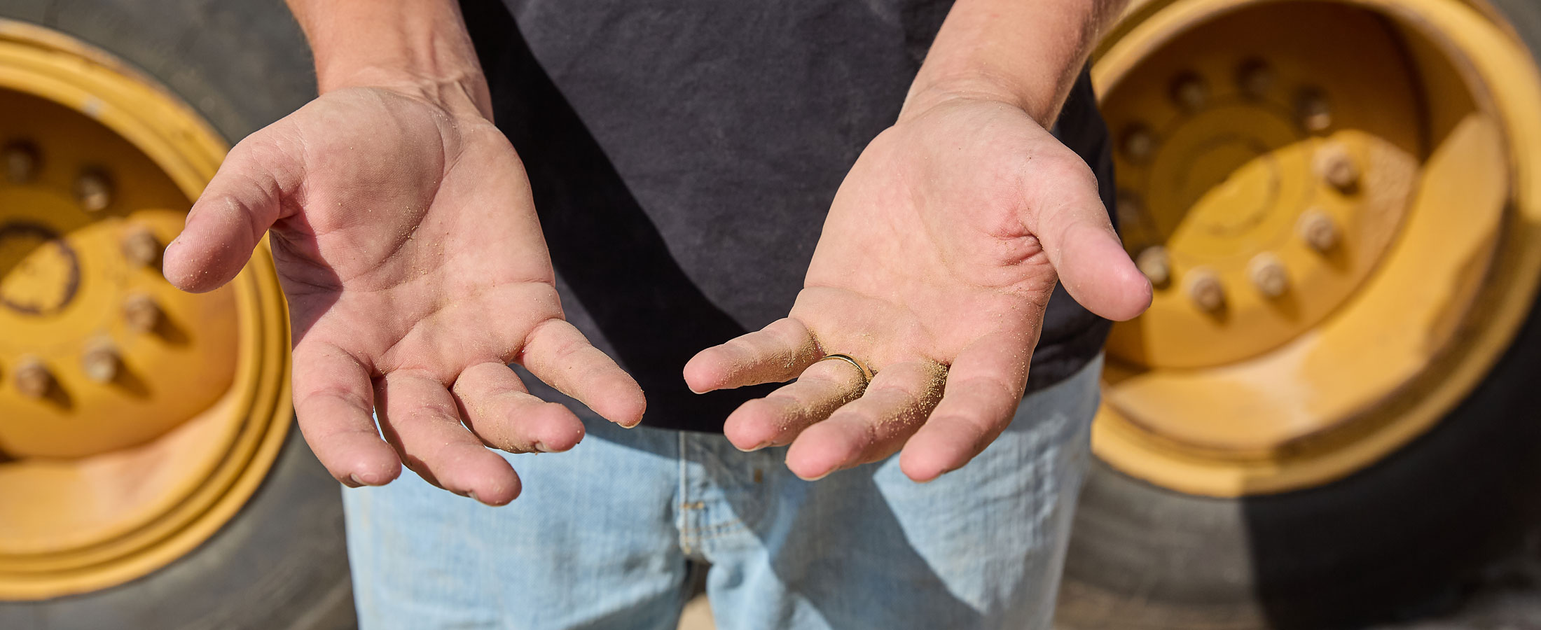 Close-up of Nick Castillion’s open hands, slightly dirty, held out in front of a large construction vehicle tire."